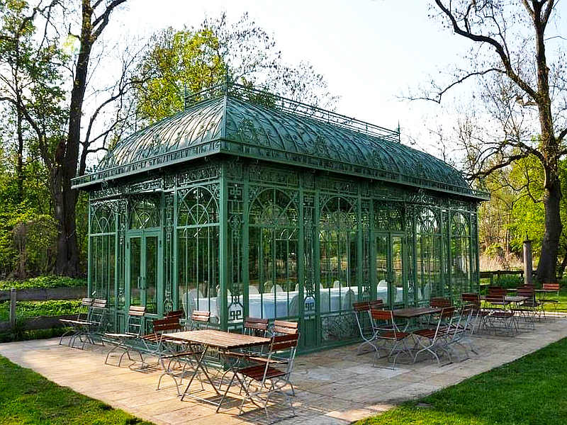 Vintage green steel Victorian greenhouse sits on stone patio among mature trees