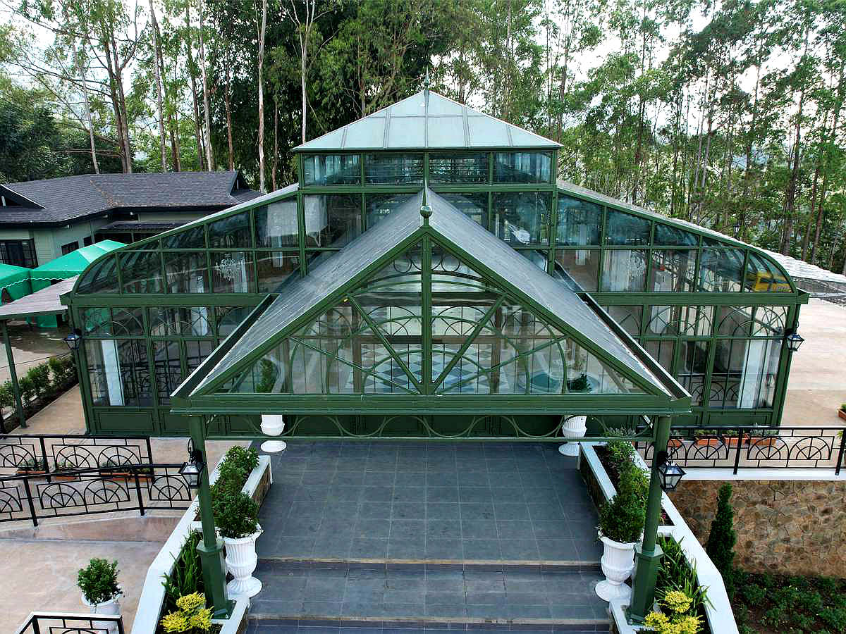Elegant green-framed glass conservatory with chandeliers, set among trees.