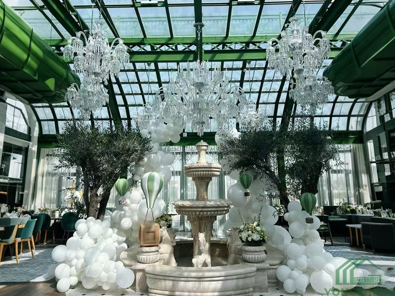 Inside Victorian glass greenhouse conservatory, high arched glass ceiling, crystal chandeliers, central stone fountain, decorated for wedding event