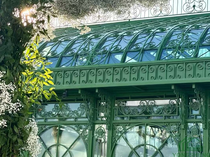 Close up of ornate green cast iron scrollwork frame on Victorian glass conservatory, arched curved glass roof detail with greenery