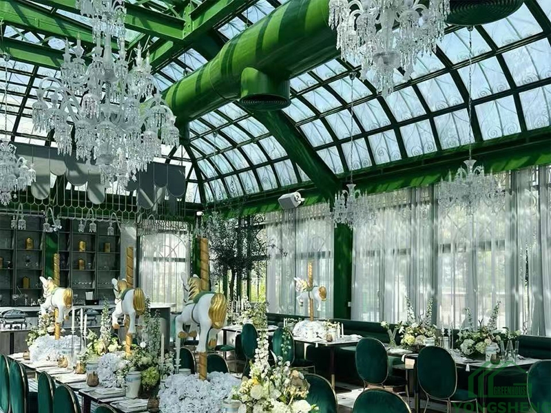 Wedding banquet long tables inside Victorian glass greenhouse, arched glass roof, hanging crystal chandeliers, green velvet dining chairs and floral decor