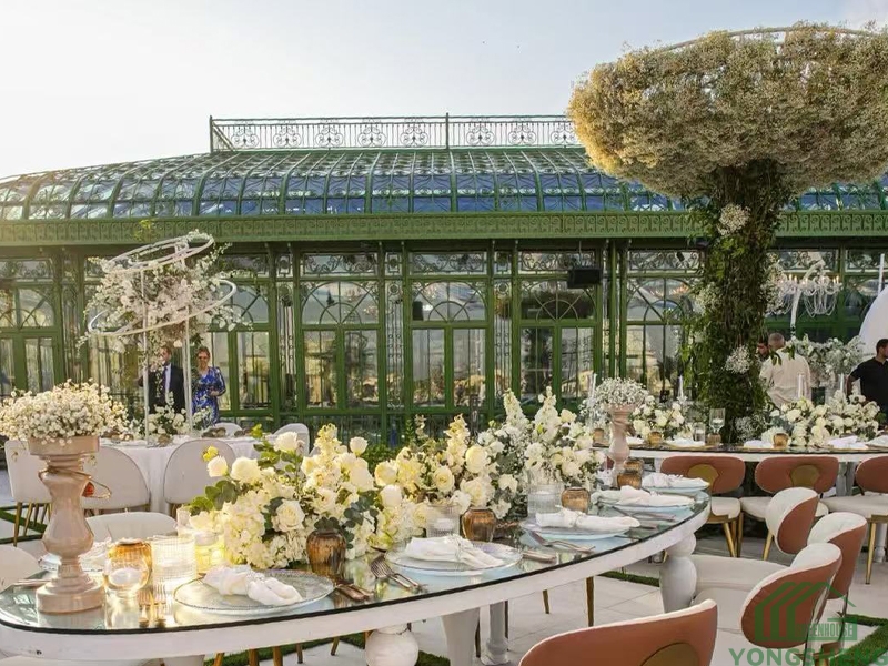 Curved glass banquet dining tables decorated with white flowers, Victorian green glass conservatory backdrop, outdoor wedding reception setup