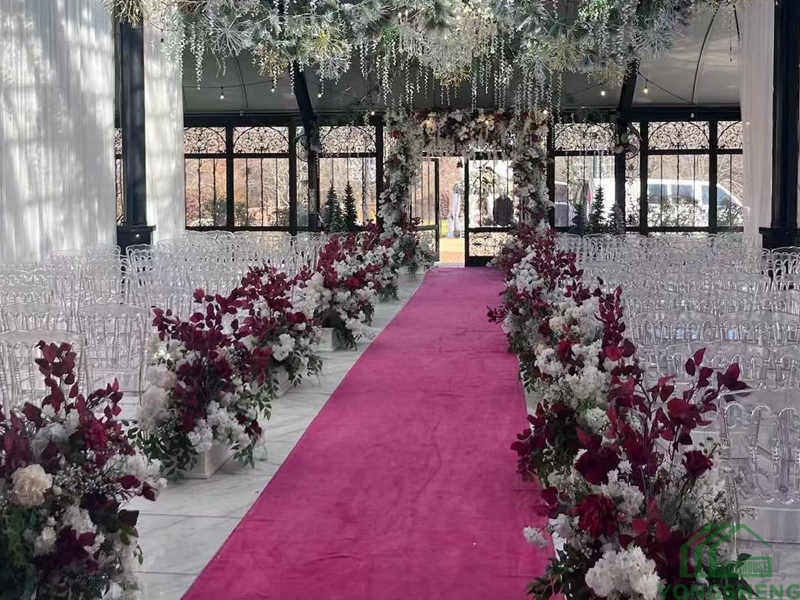 Wedding aisle with red carpet, colorful floral borders, and hanging floral decor in a glass venue.