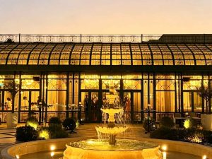 Victorian glass conservatory exterior at dusk, luxury wedding event greenhouse with curved glass roof and stone fountain in foreground, ornate green cast iron framework, warm interior lighting