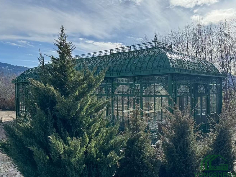 Side profile of Victorian greenhouse YSGH‑057, green steel orangerie among evergreen shrubs with mountain background