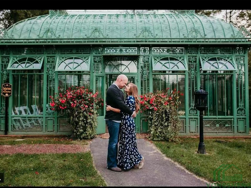 Vintage victorian green steel glass conservatory with ornate lattice arched roof, couple taking portrait photos outside