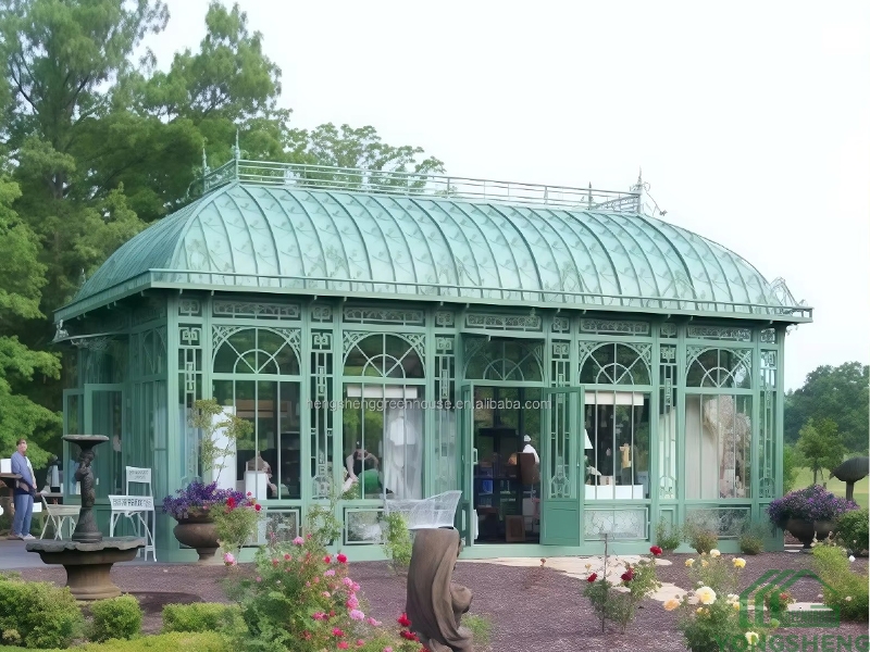 Large green victorian steel conservatory sitting in landscaped ornamental garden with flower beds and stone fountain