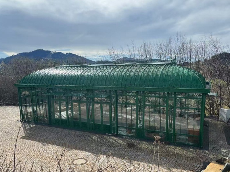 High‑angle overlook of YSGH‑057 Victorian greenhouse, long curved green steel orangerie on paved terrace