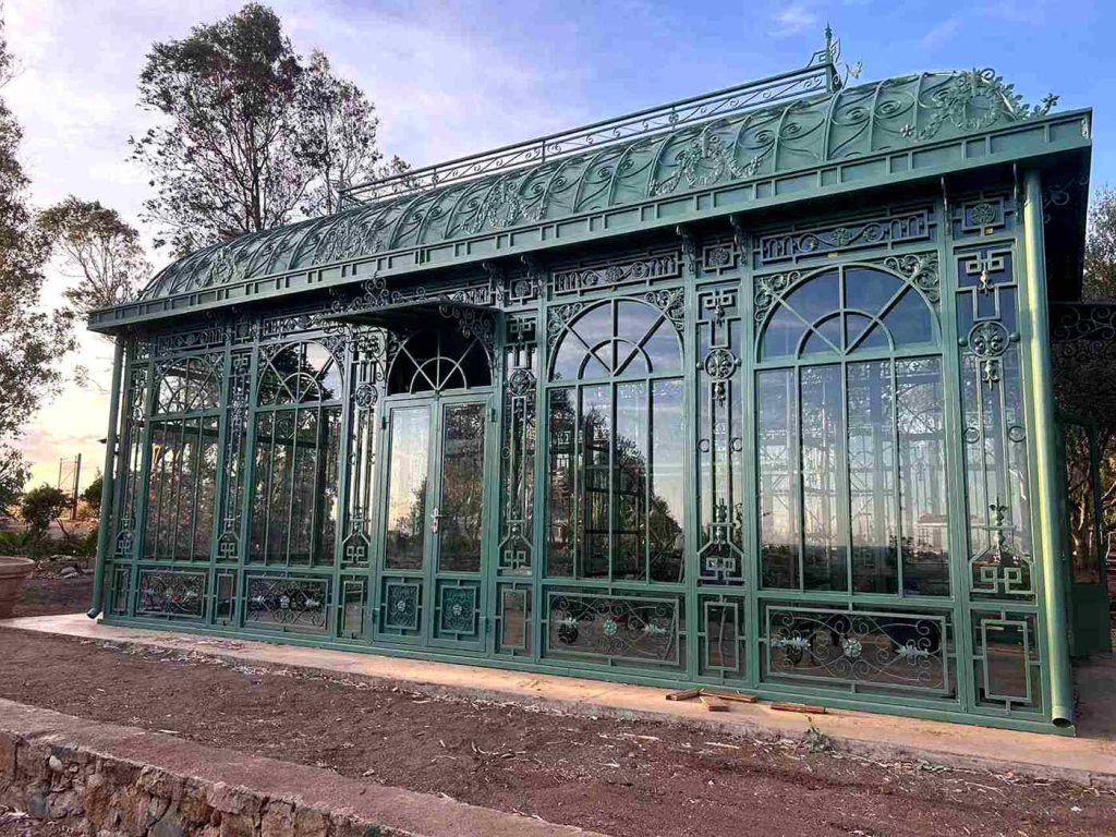 Close-up of Victorian greenhouse metal frame, arched double glass doors, and decorative roof cresting