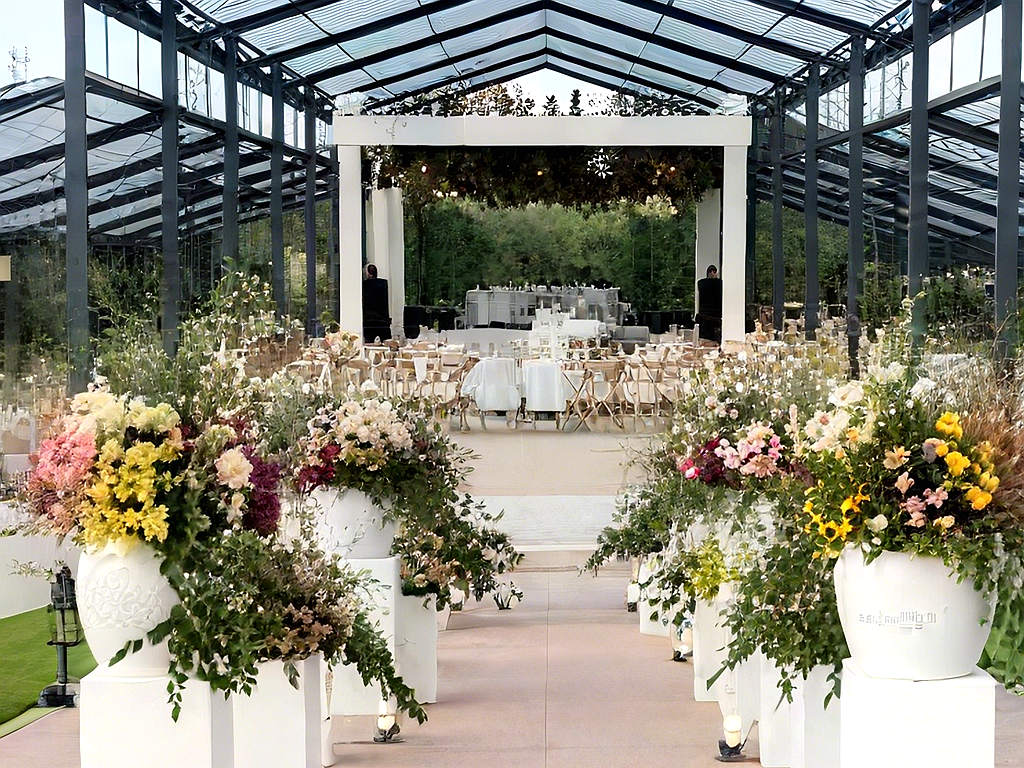 Interior view of a modern clear-roof wedding pavilion showing round banquet tables, chairs, and aisle flower pots.