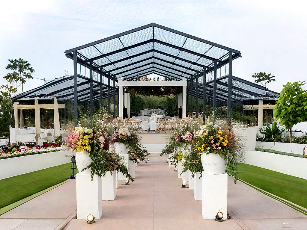Wide view of a large outdoor wedding canopy with black metal frame, clear roof, and floral decorations along the aisle.