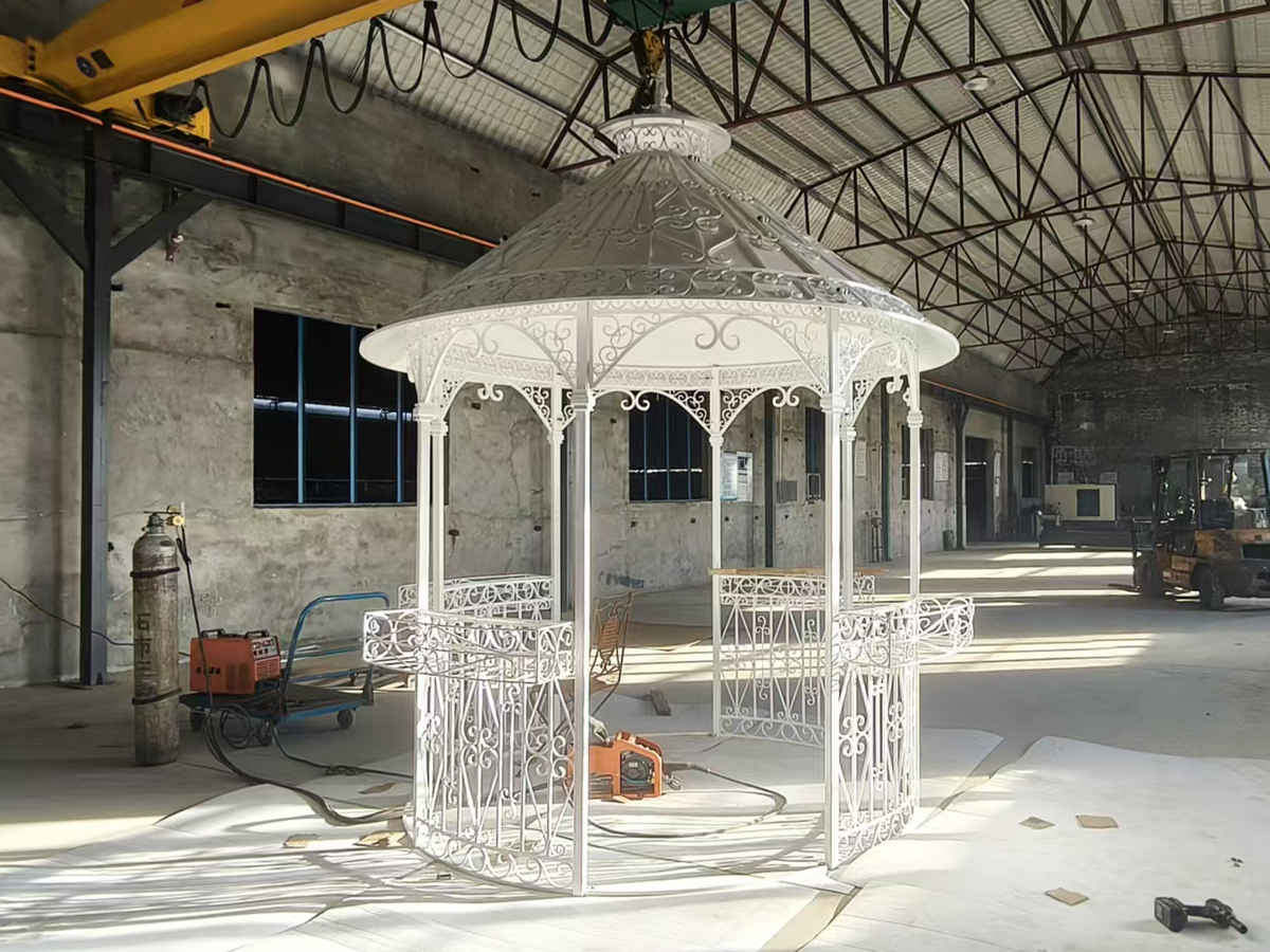 White wrought-iron octagonal gazebo being lifted by a crane in a factory workshop.