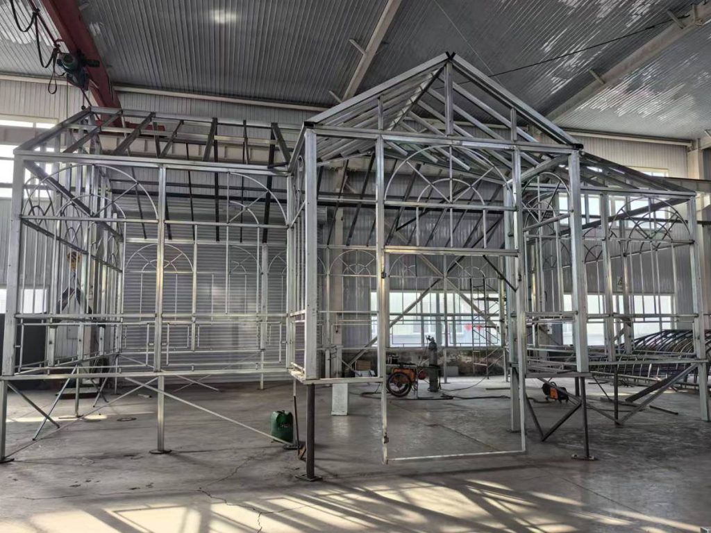 Galvanized Victorian greenhouse steel frame pre‑fabricated inside manufacturing plant, ornate arched window metal grids, gable roof conservatory framework, factory test assembly