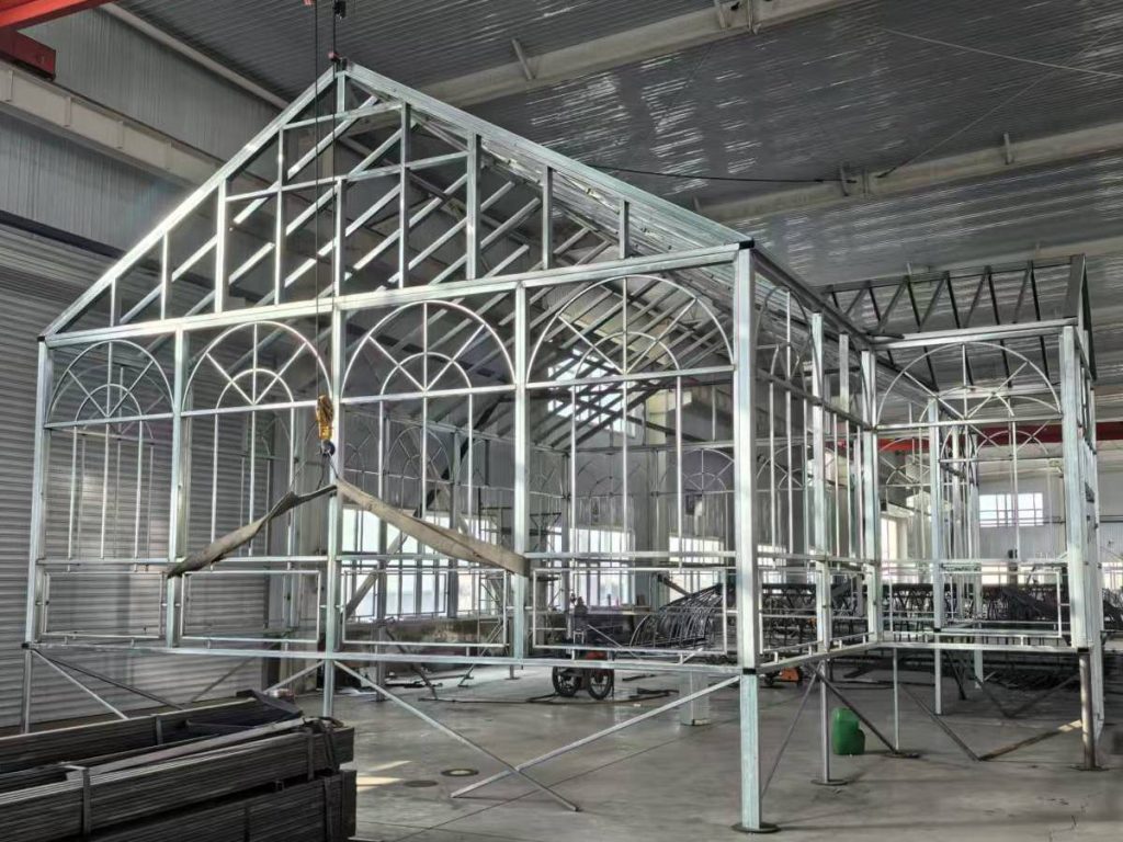 Galvanized Victorian greenhouse steel frame pre‑fabricated inside manufacturing plant, ornate arched window metal grids, gable roof conservatory framework, factory test assembly