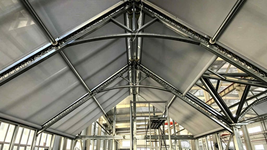 Motorized blind track system installed on arched steel roof trusses inside factory.