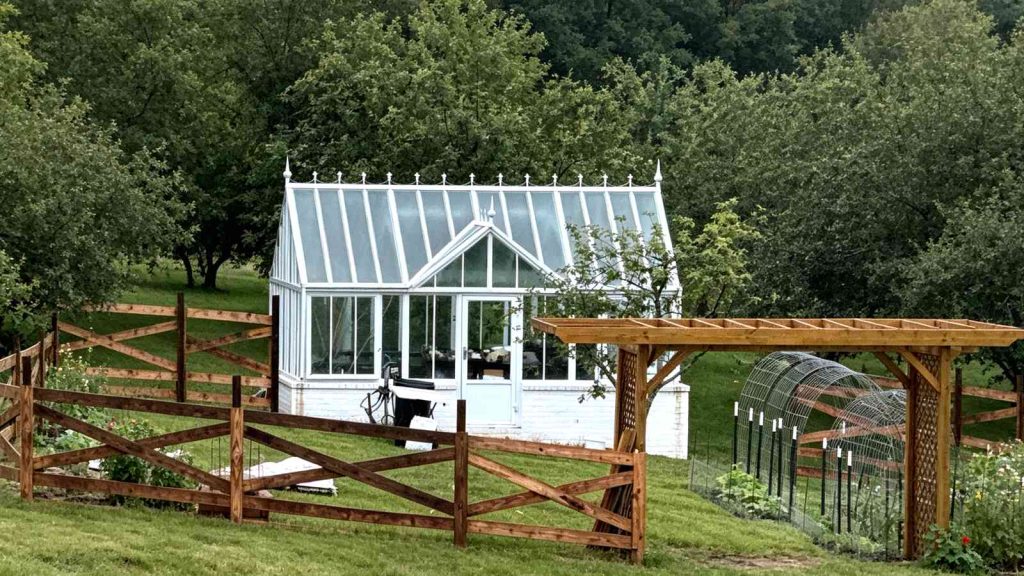 Wide shot of english‑style greenhouse surrounded by wooden fence and lush green backyard landscape