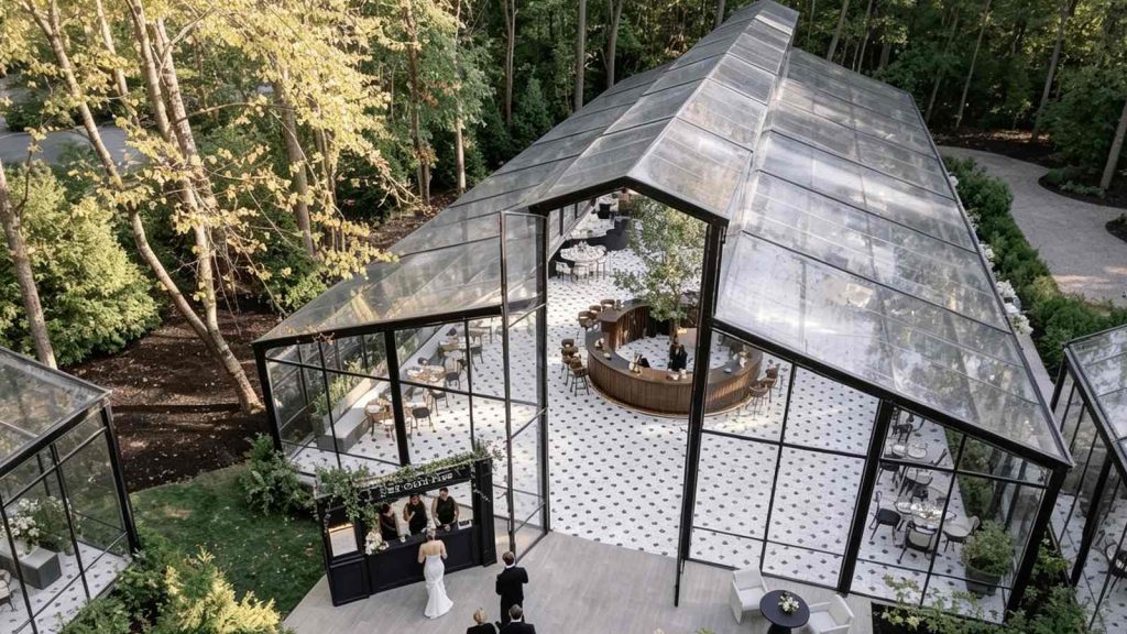 Aerial view of large wedding glass conservatory in woodland, open entrance with wedding guests and curved bar inside